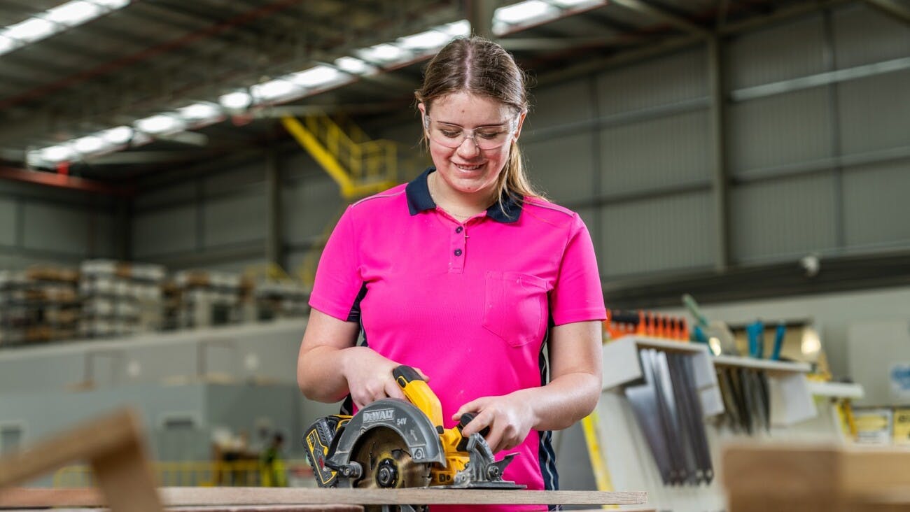Girl wearing pink shirt and safety google operating a buzz saw on a plank of wood